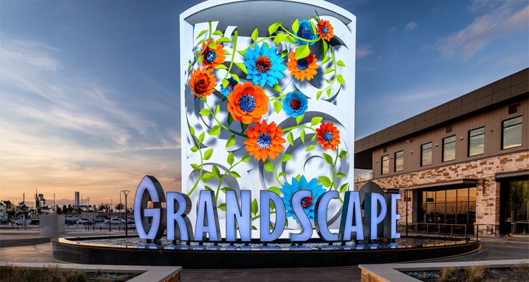 Monument sign at Grandscape in The Colony, Texas, featuring large illuminated letters spelling “GRANDSCAPE” in front of a vibrant digital display with colorful 3D floral artwork, set against a modern building and a sunset sky.