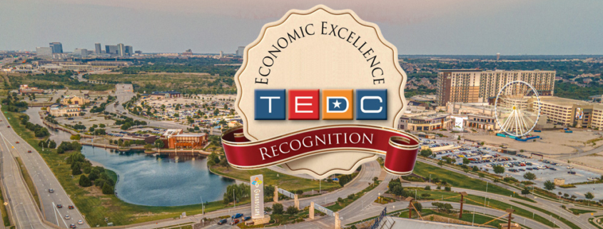 Aerial view of a Grandscape commercial district with roads, a lake, and the large Ferris wheel, overlaid with a badge reading “Economic Excellence – TEDC Recognition.”