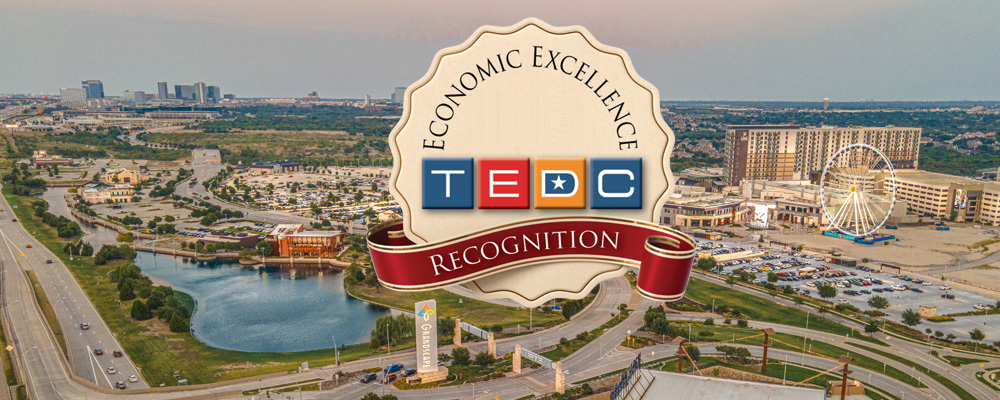 Aerial view of a Grandscape commercial district with roads, a lake, and the large Ferris wheel, overlaid with a badge reading “Economic Excellence – TEDC Recognition.”
