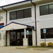 Two-story commercial office building with a light stone exterior, dark-framed windows, covered entrance, accessible parking spaces, landscaping, and tenant signage including Farmers Insurance and AmeriDream.