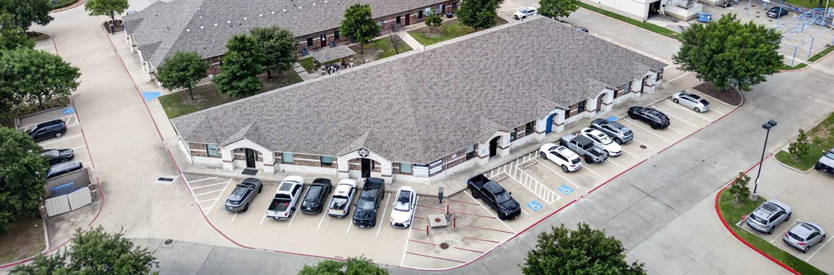 Aerial view of a single-story commercial office building with a large gray roof, surrounding parking areas, accessible parking spaces, parked vehicles, mature trees, and neighboring buildings.