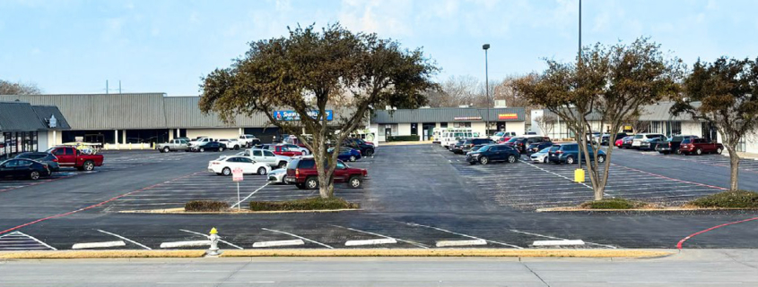 Wide view of a neighborhood shopping center and parking lot with several storefronts, parked vehicles, mature trees, accessible parking spaces, and a roadway in the foreground.