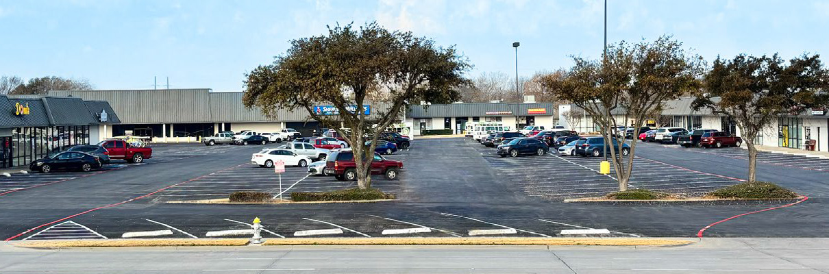 Wide view of a neighborhood shopping center and parking lot with several storefronts, parked vehicles, mature trees, accessible parking spaces, and a roadway in the foreground.