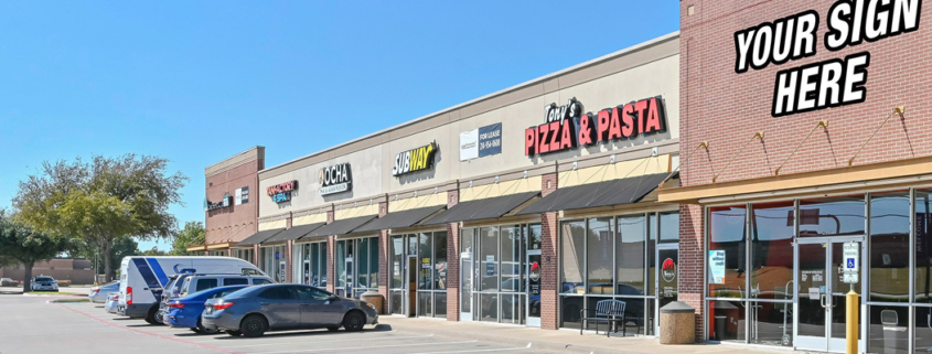 Street-level view of a retail shopping center with multiple storefronts, including restaurants and services, parked vehicles, and a prominent corner space displaying a “Your Sign Here” leasing graphic.