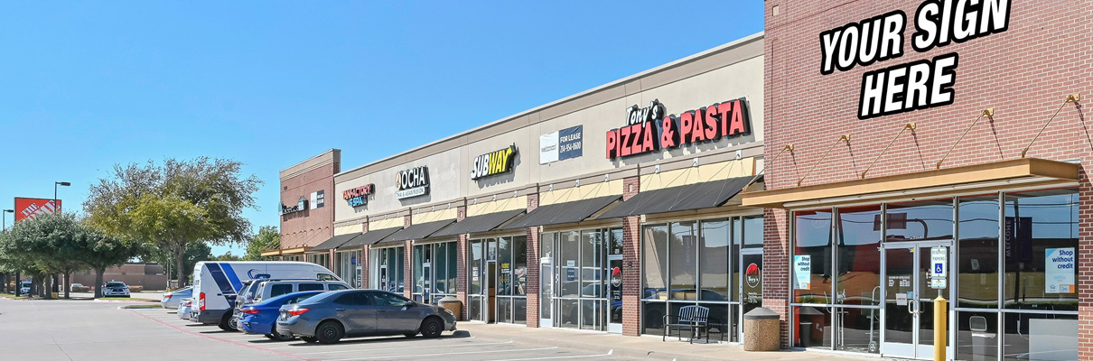 Street-level view of a retail shopping center with multiple storefronts, including restaurants and services, parked vehicles, and a prominent corner space displaying a “Your Sign Here” leasing graphic.