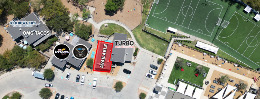 Aerial view of a mixed-use entertainment and dining area with an available commercial space highlighted in red beside Turbo Coffee & Creative, near OMG Tacos, The Empanada Cookhouse, outdoor gathering areas, and adjacent soccer fields.