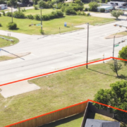 Aerial view of a vacant commercial property outlined in red at a signalized intersection, with surrounding roads, businesses, parking areas, trees, and nearby development.
