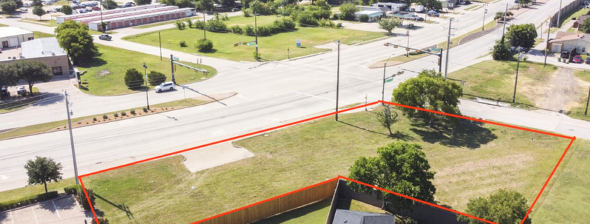 Aerial view of a vacant commercial property outlined in red at a signalized intersection, with surrounding roads, businesses, parking areas, trees, and nearby development.