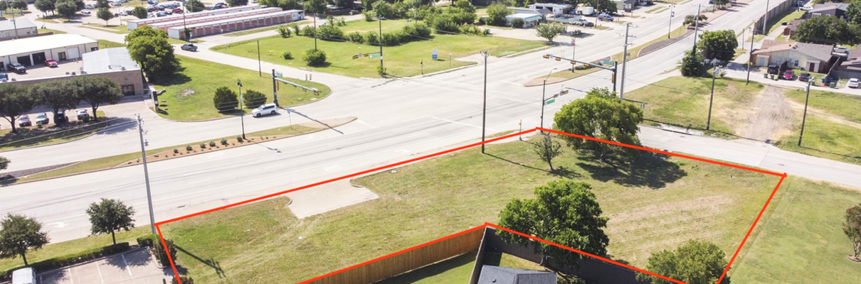 Aerial view of a vacant commercial property outlined in red at a signalized intersection, with surrounding roads, businesses, parking areas, trees, and nearby development.