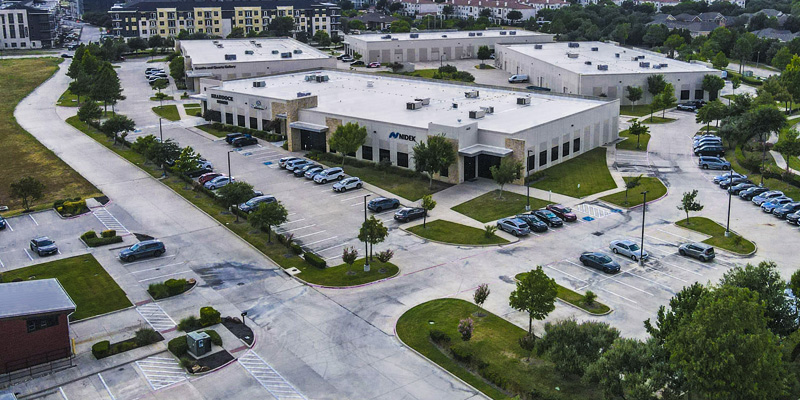 Aerial view of a commercial office complex with multiple buildings, landscaped parking lots, parked vehicles, and nearby residential development.